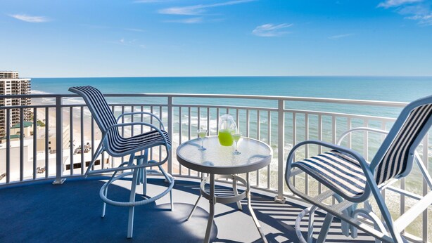A balcony with two striped chairs and a small round table, overlooking a clear blue sea and distant buildings.