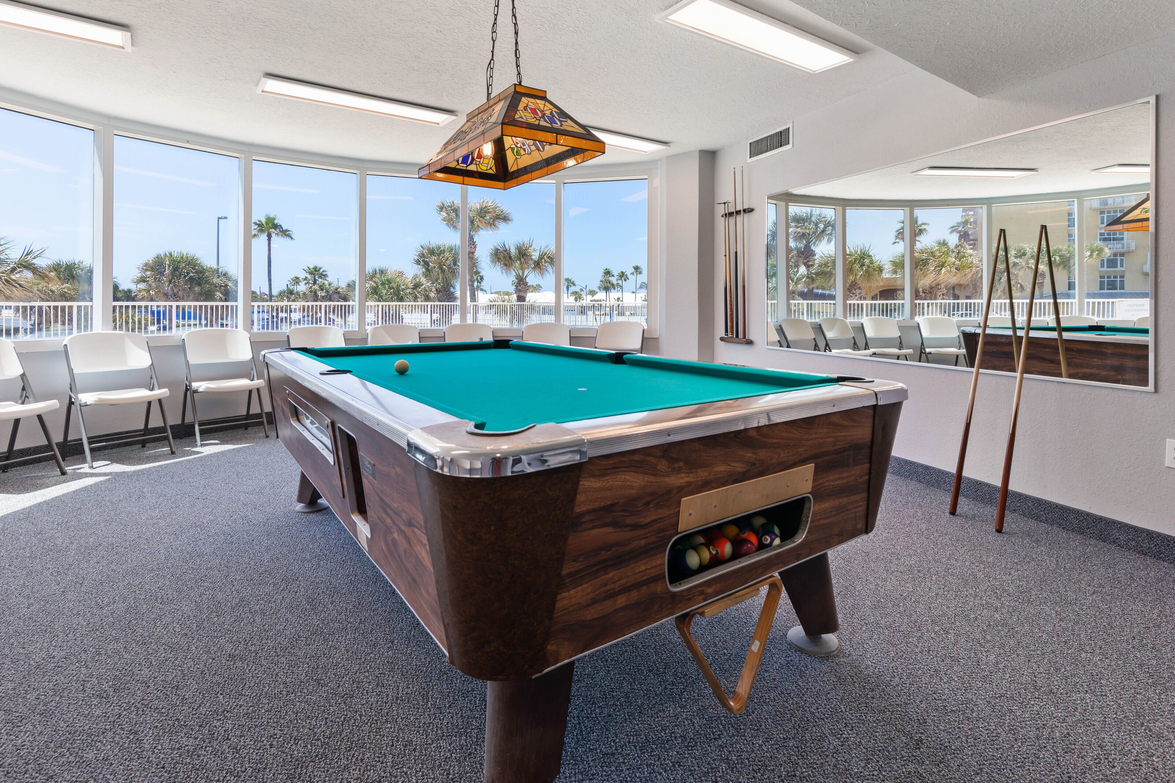 A pool table with a wooden finish and a hanging light fixture. There are chairs around the table and a large window with a view of palm trees.