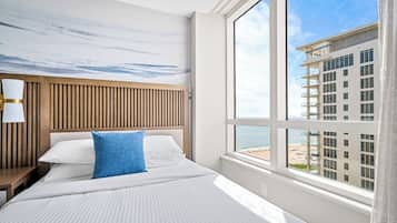 Ein Hotelzimmer mit großem Fenster, einem Holzkopfteil und Blick auf Strand und Häuser.