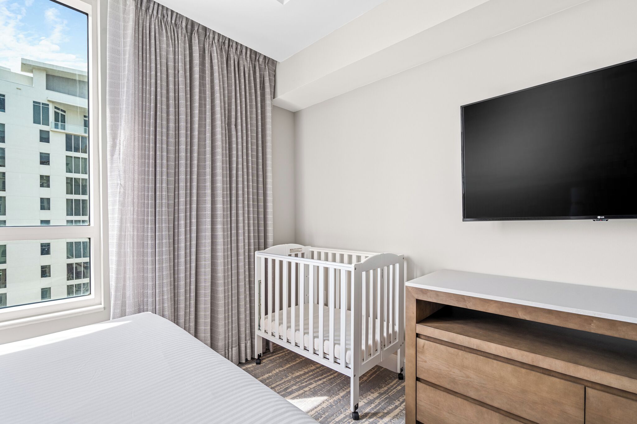 A hotel room with a crib, a TV, and a large window with curtains.