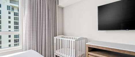 A hotel room with a crib, a TV, and a large window with curtains.