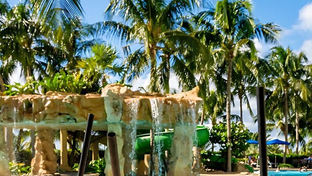 A poolside view with three drinks in front of a water slide and palm trees. - Singer Island Palm Beach Marriott: 2 BDRM/2 BATH Condo +Resort Access 14th Floor (Riviera Beach)
