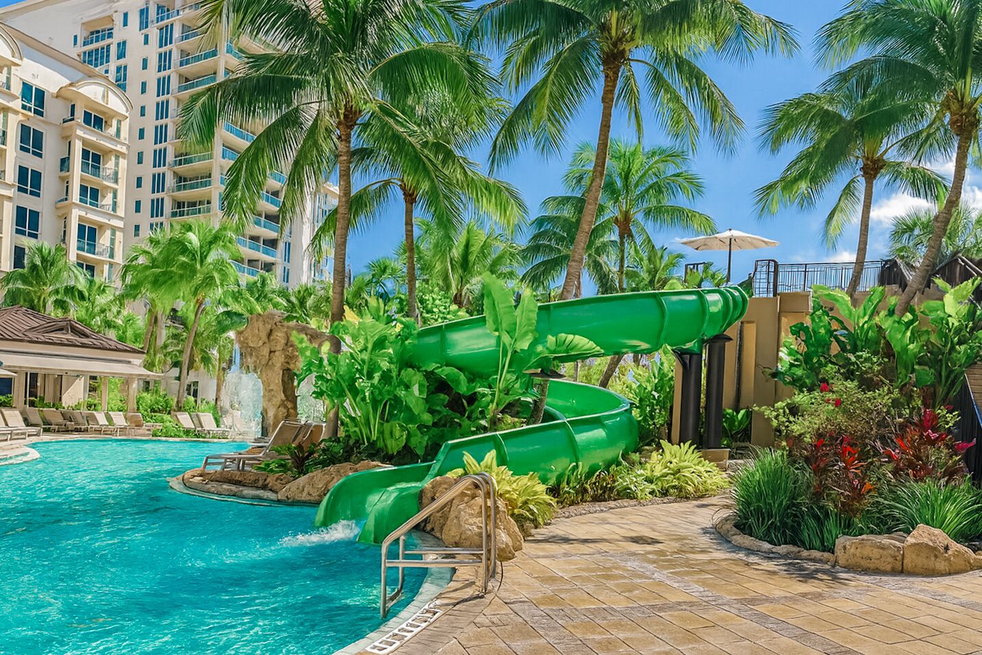 A hotel pool area with a water slide, palm trees, and a clear blue sky.
