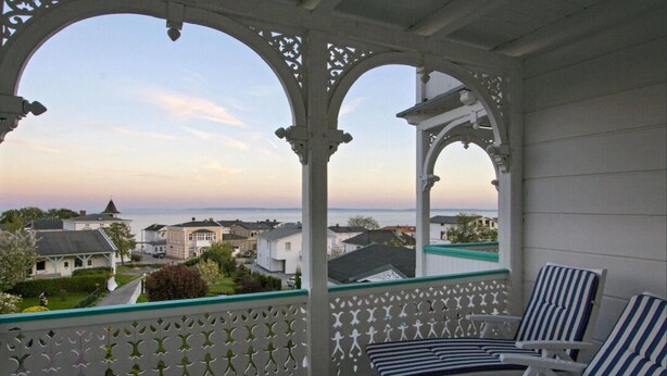 Ein überdachter Balkon mit weißen Gittergeländern und blau gestreiften Liegestühlen, mit Blick auf eine Wohngegend und ein Gewässer.