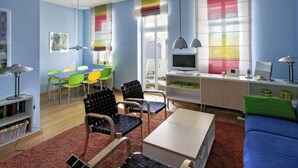 Ein modernes Wohnzimmer mit einem blauen Sofa, einem Couchtisch und einem Fernseher. Bunte Sessel und ein Esstisch mit Stühlen sind ebenfalls vorhanden.