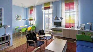 Ein modernes Wohnzimmer mit einem blauen Sofa, einem Couchtisch und einem Fernseher. Bunte Sessel und ein Esstisch mit Stühlen sind ebenfalls vorhanden.