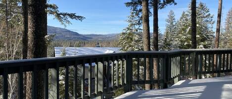 A snow-covered deck with a railing overlooking a forested area and distant mountains.