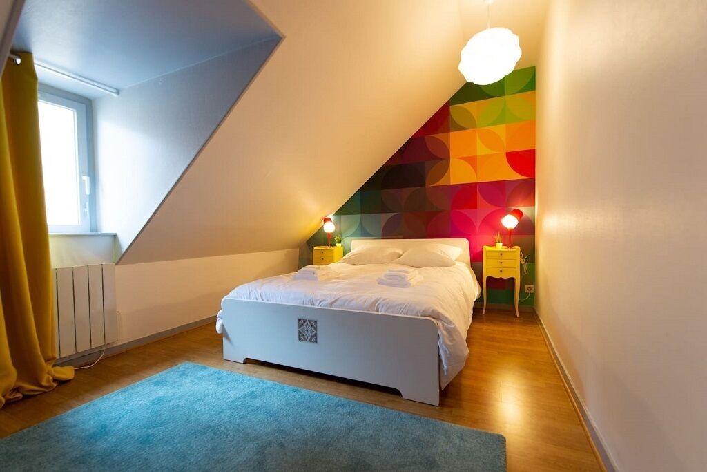 A bedroom with a sloped ceiling, a bed with white bedding, two bedside tables with lamps, and a colorful geometric wall design.