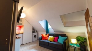Un espacio habitable compacto con kitchenette, un sofá con cojines coloridos, una mesita con lámpara y una planta en maceta.