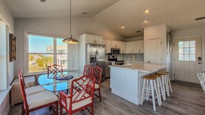 A modern kitchen with white cabinets, a stainless steel refrigerator, a microwave, and a built-in oven. There is a dining area with a glass table and red chairs.