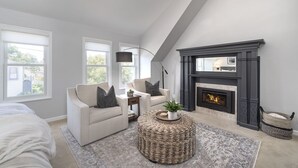 A modern living room with a fireplace, two sofas, a round wicker ottoman, and a large window. - 3813 Point Break ~ Walk to Lover's Point Beach! (Pacific Grove)