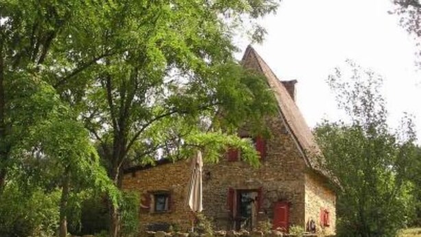 A stone house with a steeply pitched roof, surrounded by lush greenery and trees.