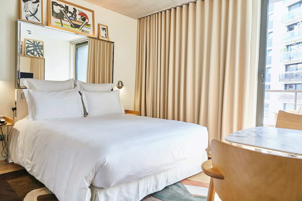 A hotel room with a large bed, a bedside table, a chair, a round table, and a window with curtains.