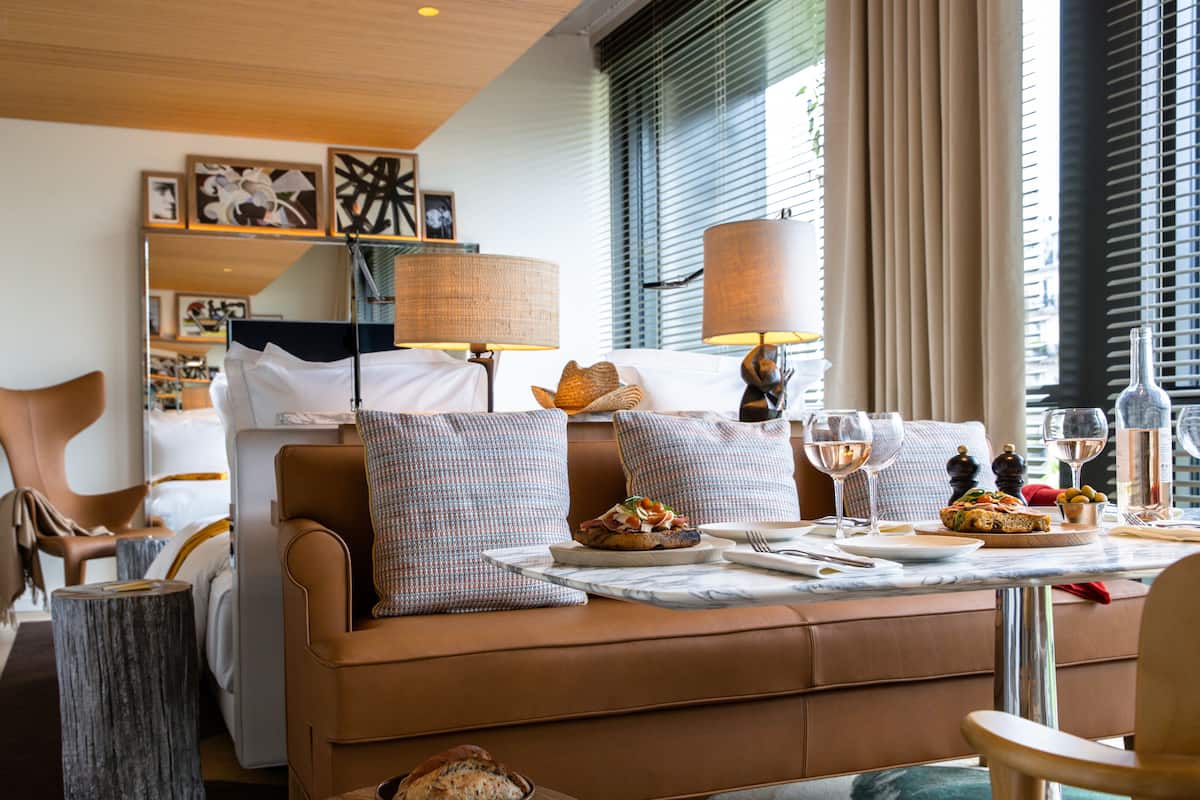 A hotel room with a sofa bed, a dining table set with plates, glasses, and a bottle. There are pillows on the bed, a lamp, and artwork on the wall.