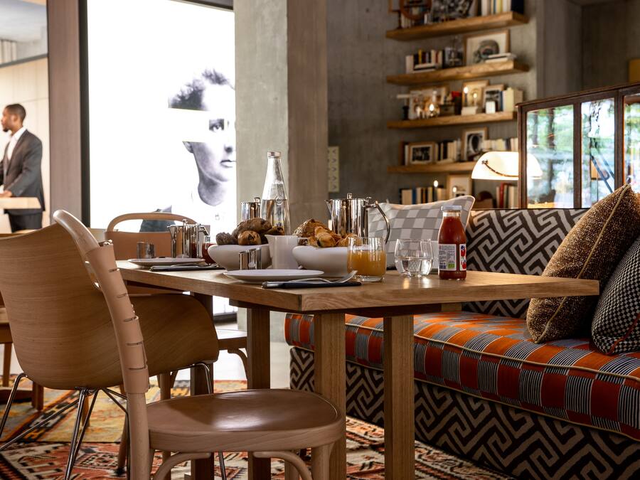 Un comedor con una mesa de madera dispuesta para el desayuno, un sofá con cojines estampados, y una estantería al fondo.