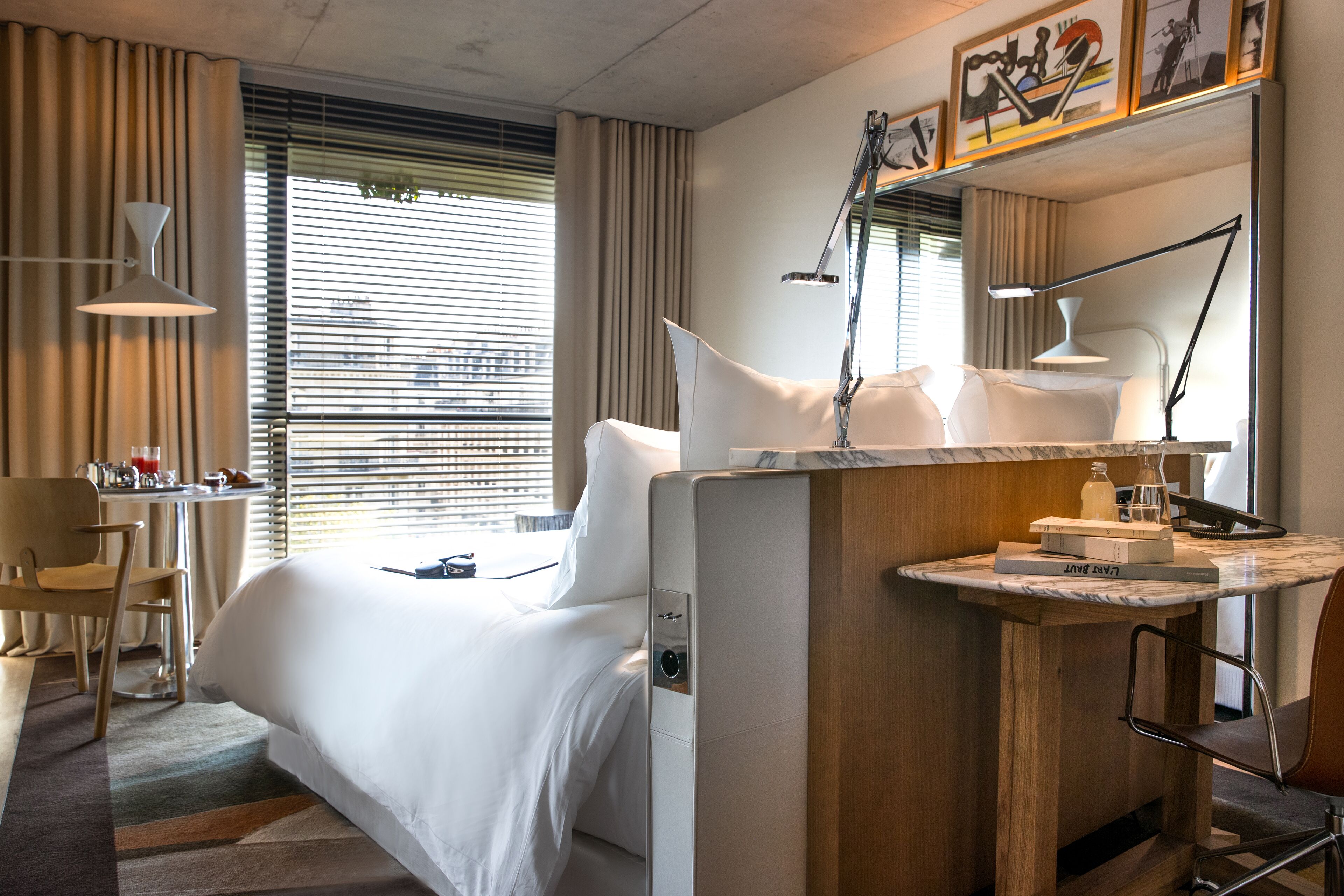 A hotel room with a bed, bedside table, lamp, and a small desk with a chair.