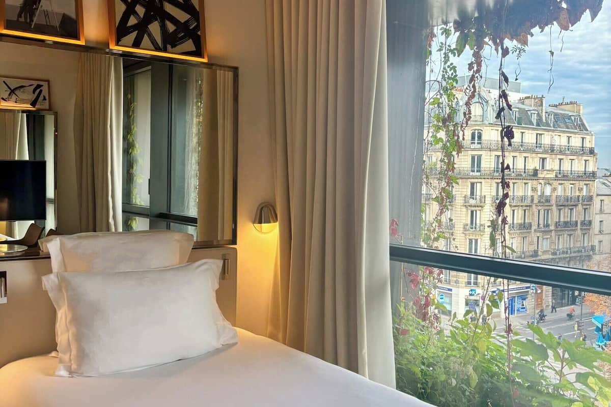A hotel room with a bed, a large window with curtains, a mirror, and framed artwork on the wall.