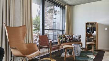 Una sala de estar moderna con un ventanal grande, un sillón de cuero marrón, un sofá de madera con cojines estampados y una pequeña mesa redonda.