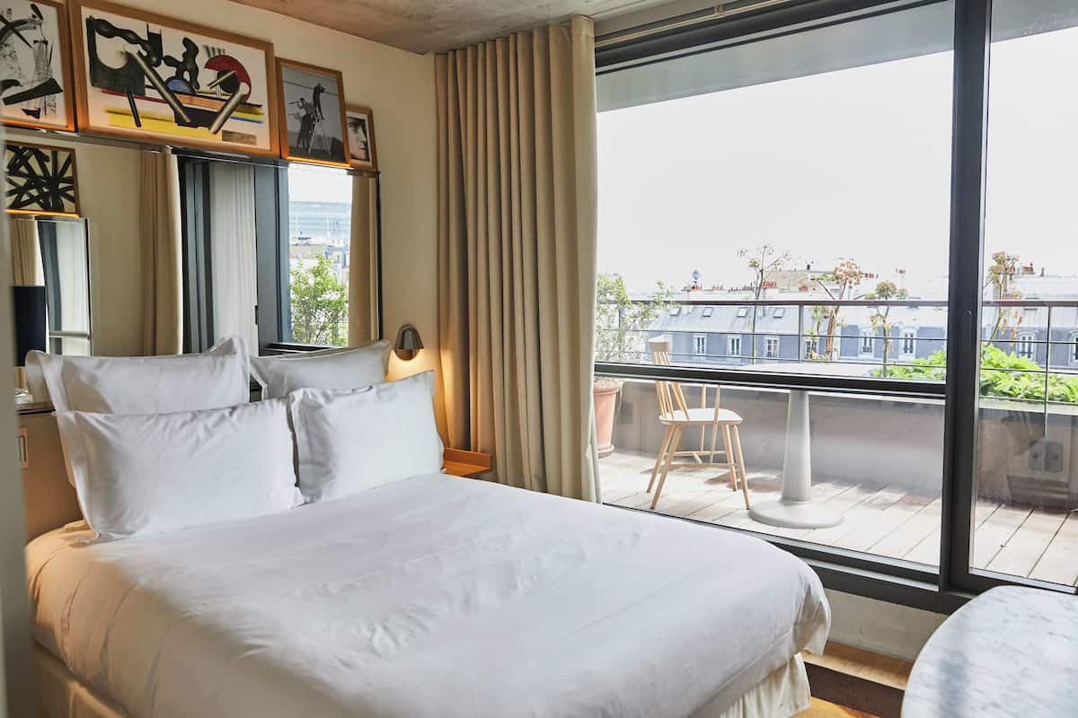 A hotel room with a large bed, a window with a view of buildings, a balcony with a chair, and a table.