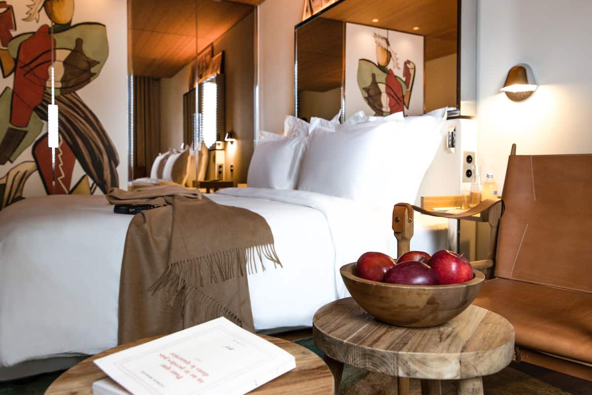 A hotel room with two beds, a wooden side table with a bowl of apples, a brown throw blanket, and a lamp.