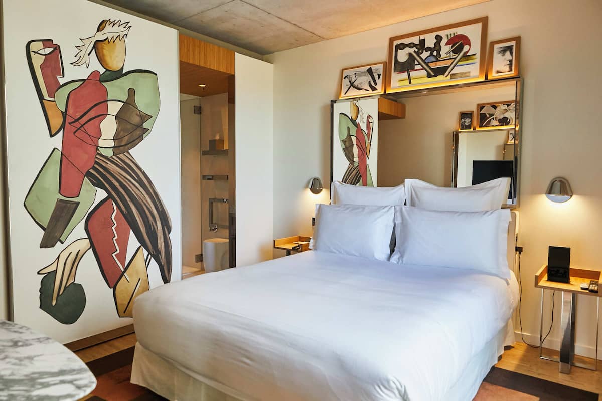 A hotel room with a large bed, a bedside table with a lamp, a wall-mounted mirror, and a painting on the wall.