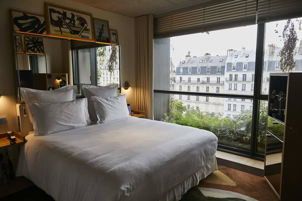 A hotel room with a large bed, a bedside table, a mirror, and a window with a view of buildings.