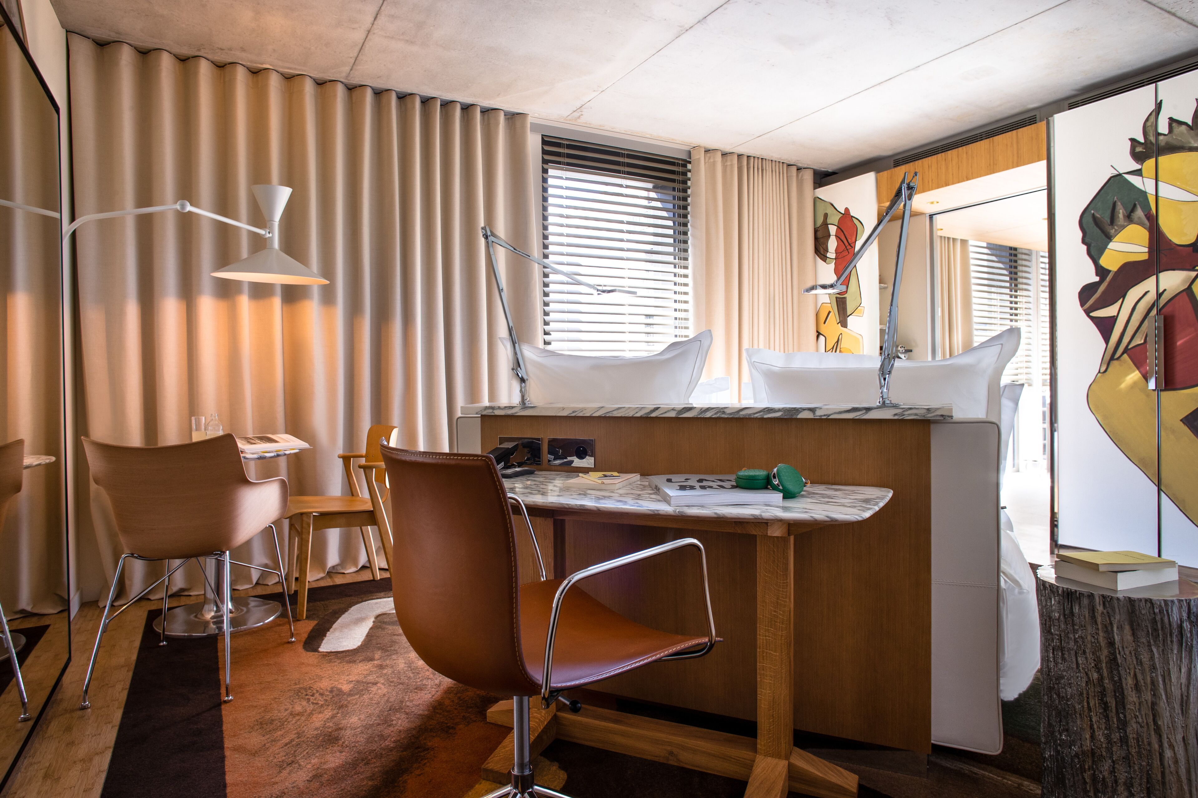 Une chambre d’hôtel moderne avec un bureau en bois, deux chaises, une petite table, un lavabo et une fenêtre avec des stores.