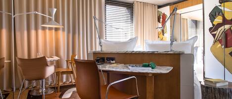 A modern hotel room with a wooden desk, two chairs, a small table, a sink, and a window with blinds.