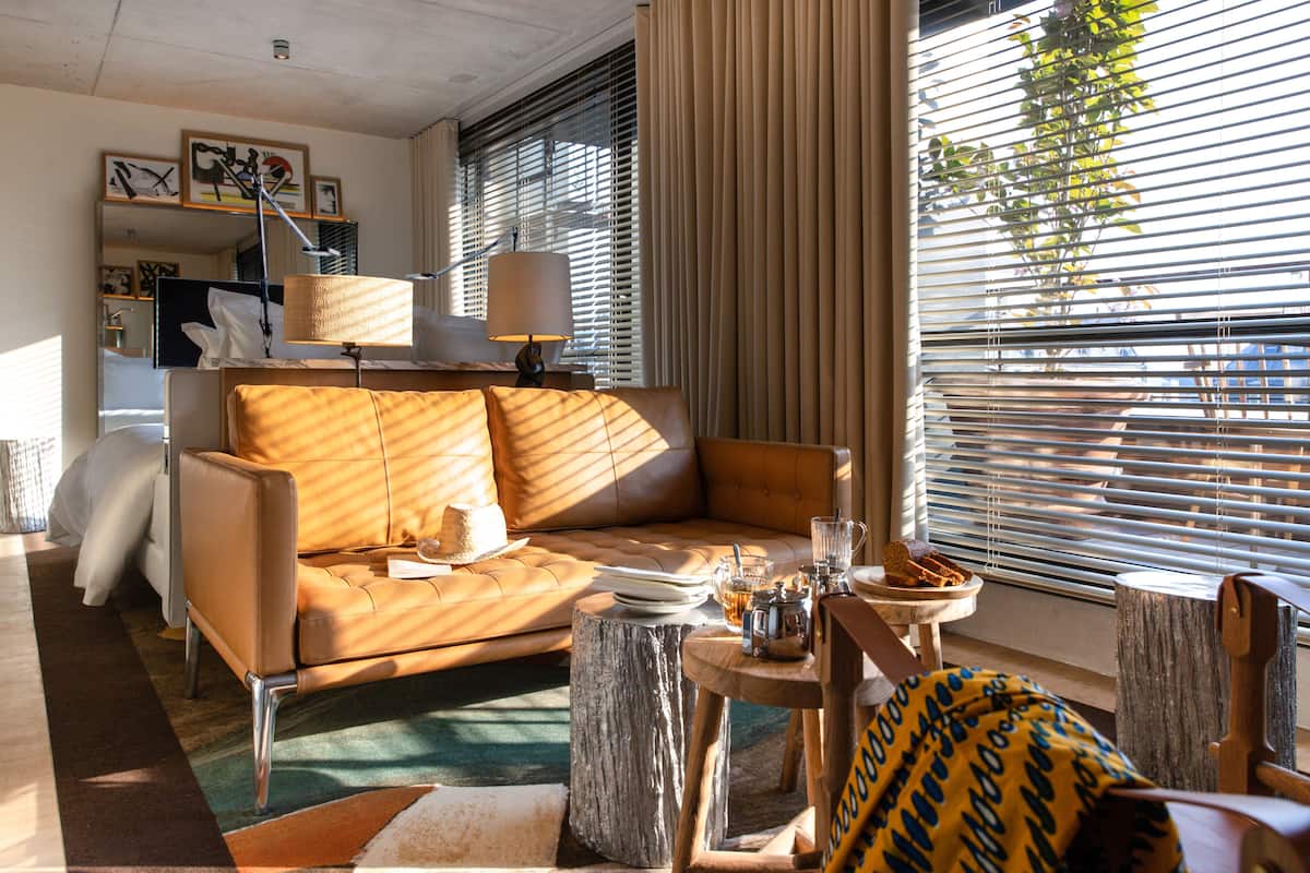 A modern hotel room with a leather sofa, a wooden side table, and a large window with blinds.