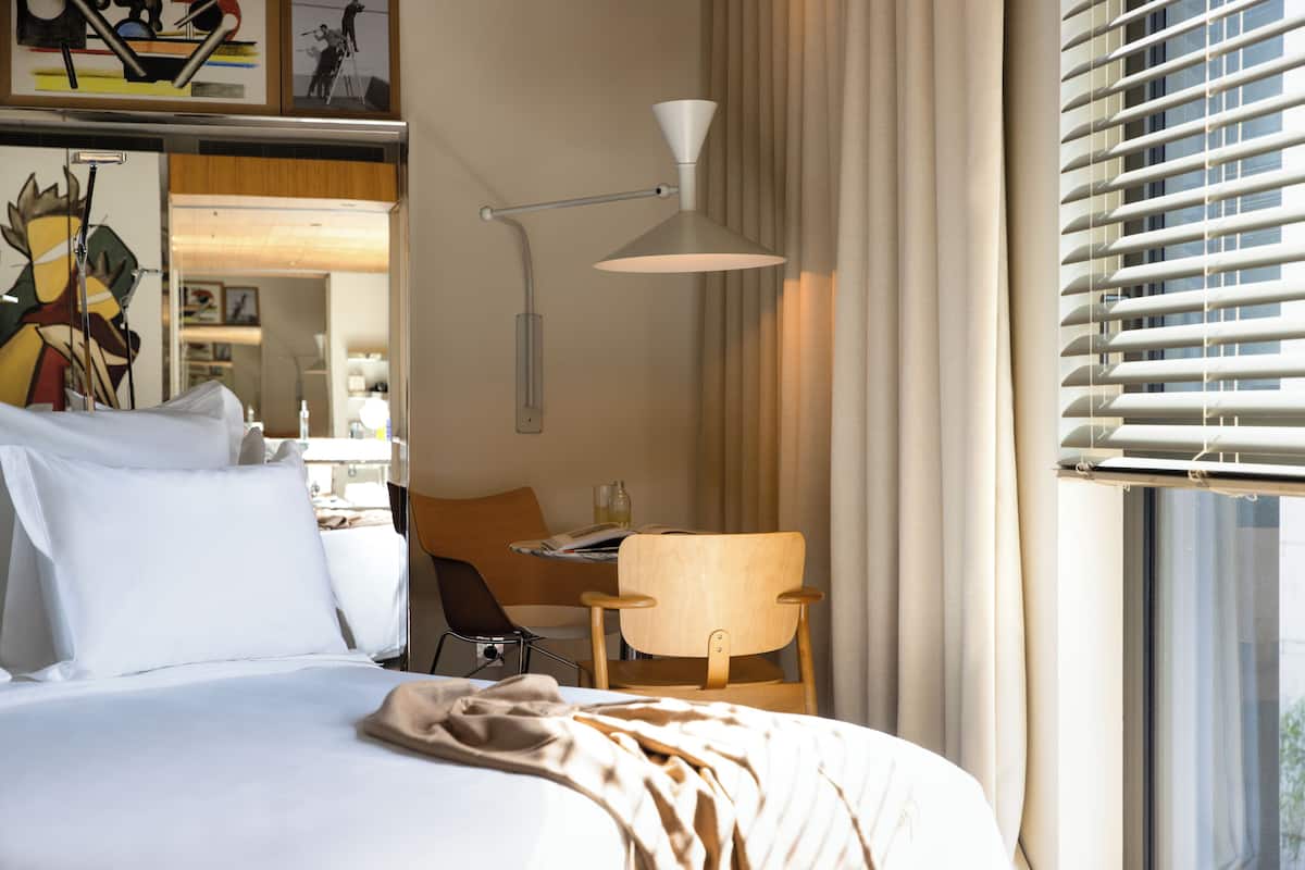 A hotel room with a bed, a desk, a chair, a lamp, and a window with blinds.