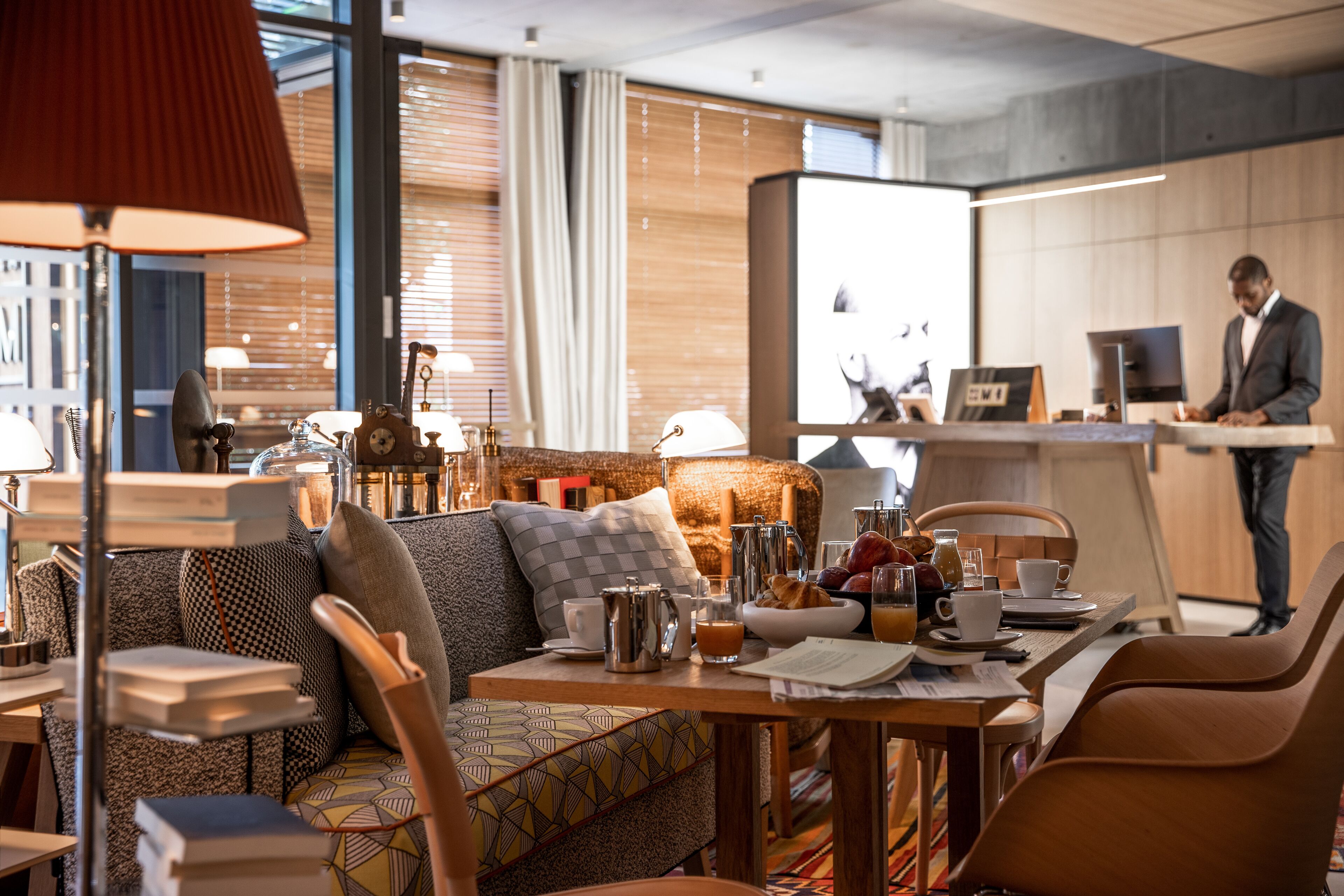 Habitación de hotel moderna con zona de comedor, sofá, mesa con artículos de desayuno y un gran ventanal con cortinas.
