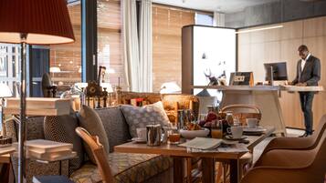 Habitación de hotel moderna con zona de comedor, sofá, mesa con artículos de desayuno y un gran ventanal con cortinas.