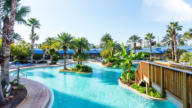 Ein Poolbereich eines Resorts mit Palmen, Liegestühlen und einer Holzbrücke.