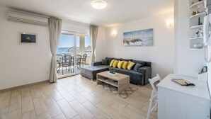 A modern living room with a grey sofa, a wooden coffee table, and a white desk with a notebook. There is a painting on the wall and a view of the outdoors through a glass door. - Charming apart.sa Terasom,250m od Mora,Parking,Wi-Fi,Roštilj,4 zvjezdice**** (Okrug Gornji)