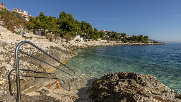 A coastal scene with clear water, rocky steps leading to a beach, and houses in the background. - Charming apart.sa Terasom,250m od Mora,Parking,Wi-Fi,Roštilj,4 zvjezdice**** (Okrug Gornji)