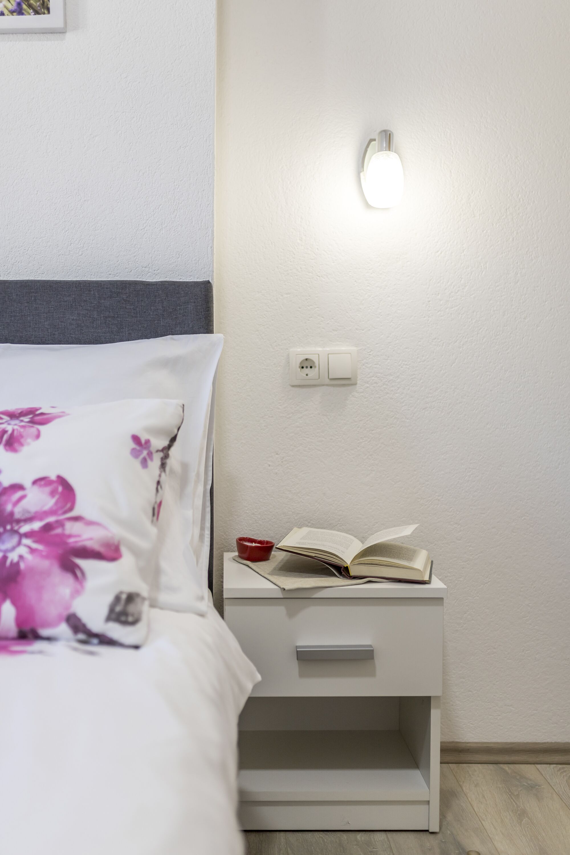 A neatly made bed with a floral pillow, a bedside table with a book and a red bowl, and a wall-mounted light fixture.
