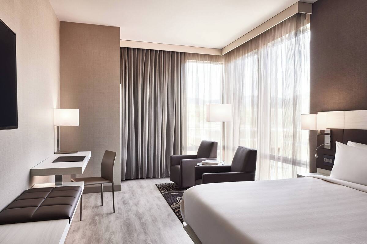 A modern hotel room with a large bed, a desk with a chair, a small table with a chair, and a sofa.