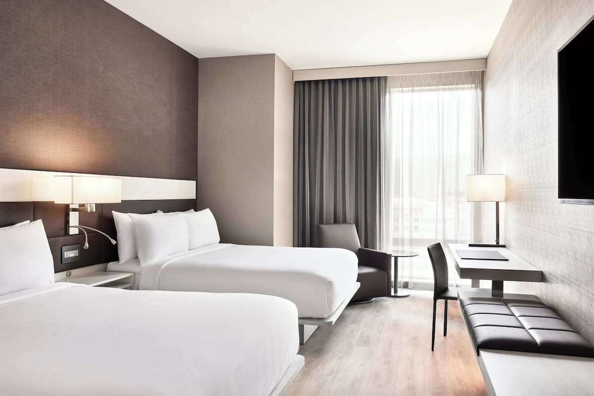 A hotel room with two beds, a desk, a chair, and a bench.