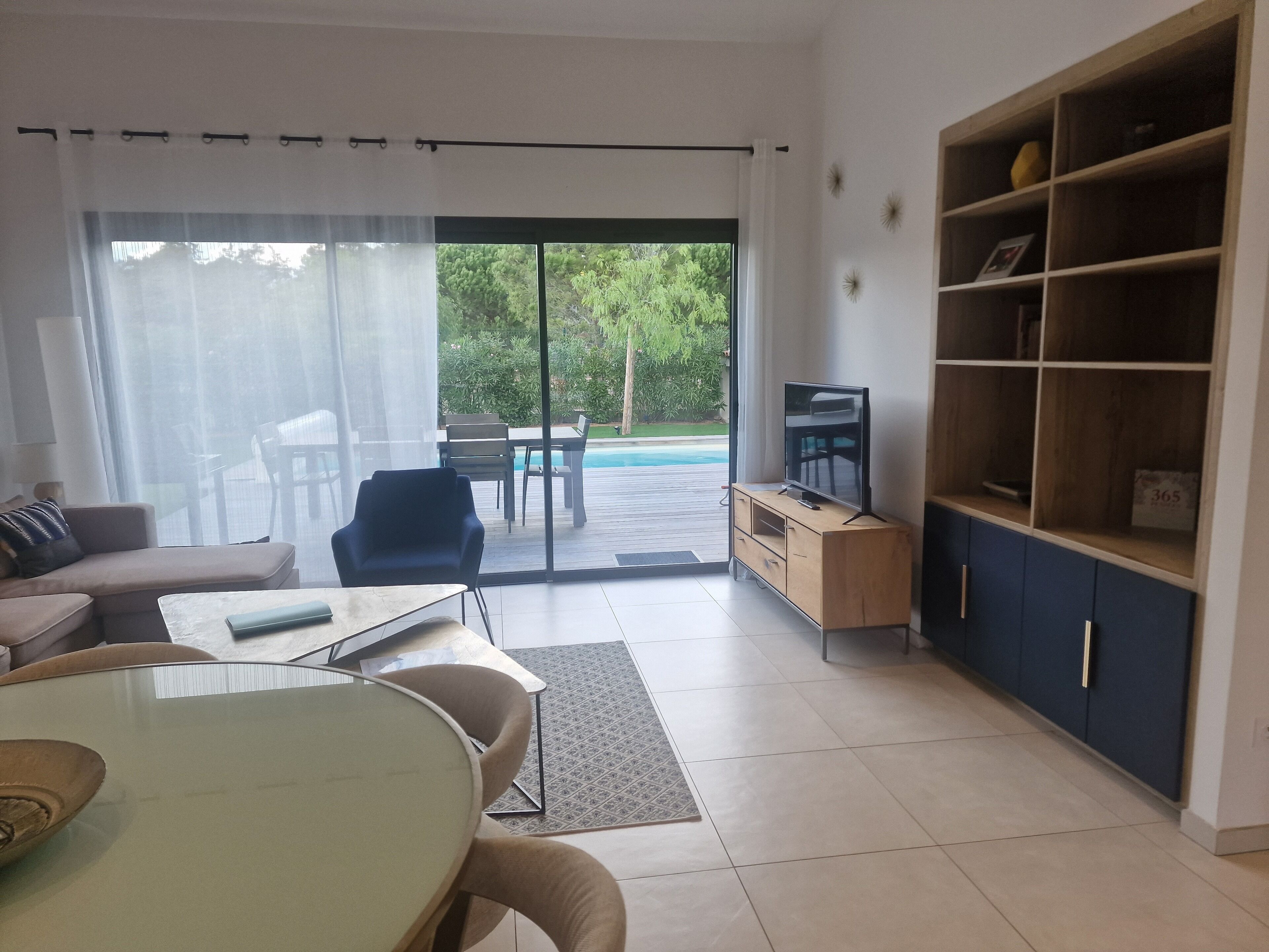 A modern living room with a sofa, a dining table, a TV on a wooden cabinet, and a view of a pool through large glass doors.