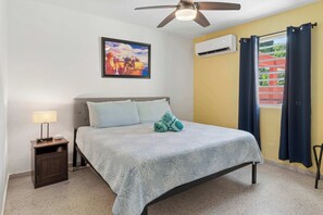 A bed with a patterned bedspread, a nightstand with a lamp, a ceiling fan, and an air conditioning unit.