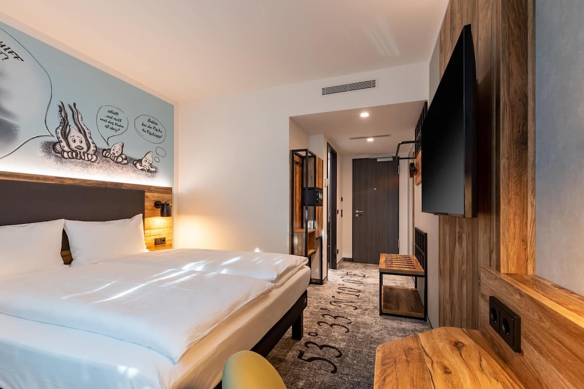 Een hotelkamer met een bed, een aan de muur bevestigde televisie, een houten bureau en een deur die leidt naar een andere kamer.