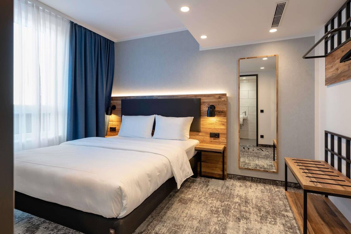 Een moderne hotelkamer met een bed, nachtkastje, spiegel en een klein houten bankje.