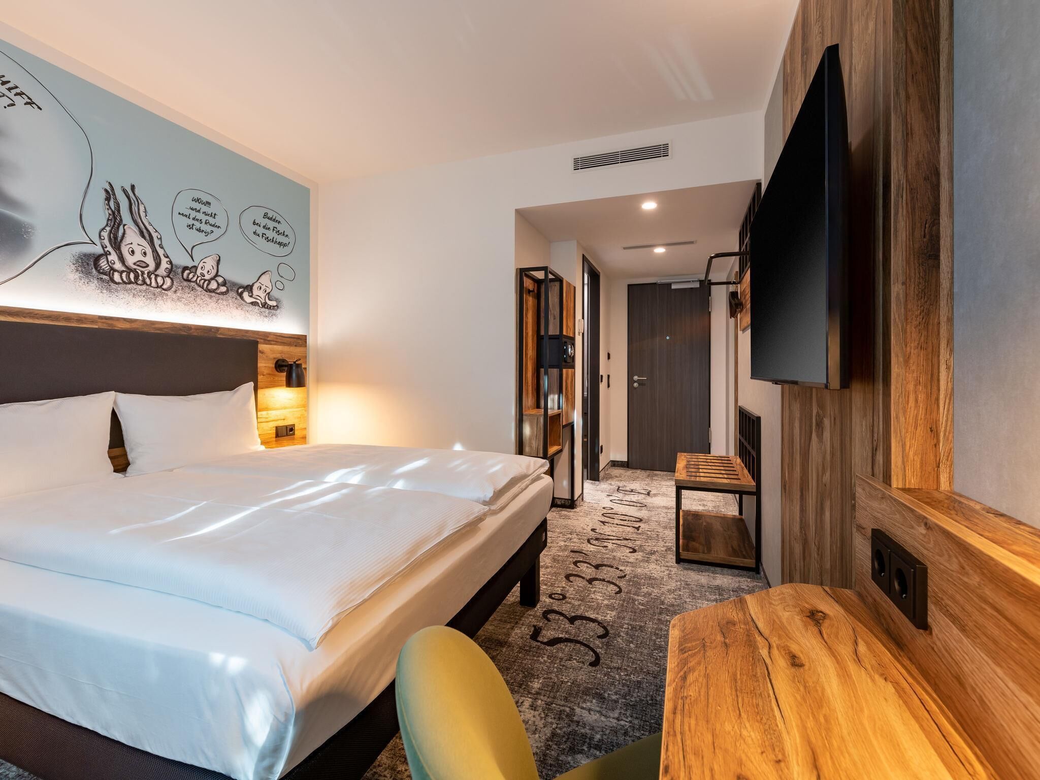 Une chambre d’hôtel équipée d’un lit, d’un bureau, d’une télévision et d’un mur décoré d’une fresque murale représentant des personnages de dessins animés.