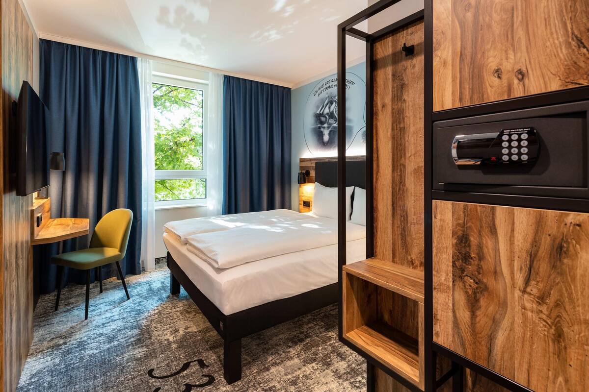 A hotel room with a bed, a desk, a chair, a TV, a wall-mounted control panel, and a window with curtains.