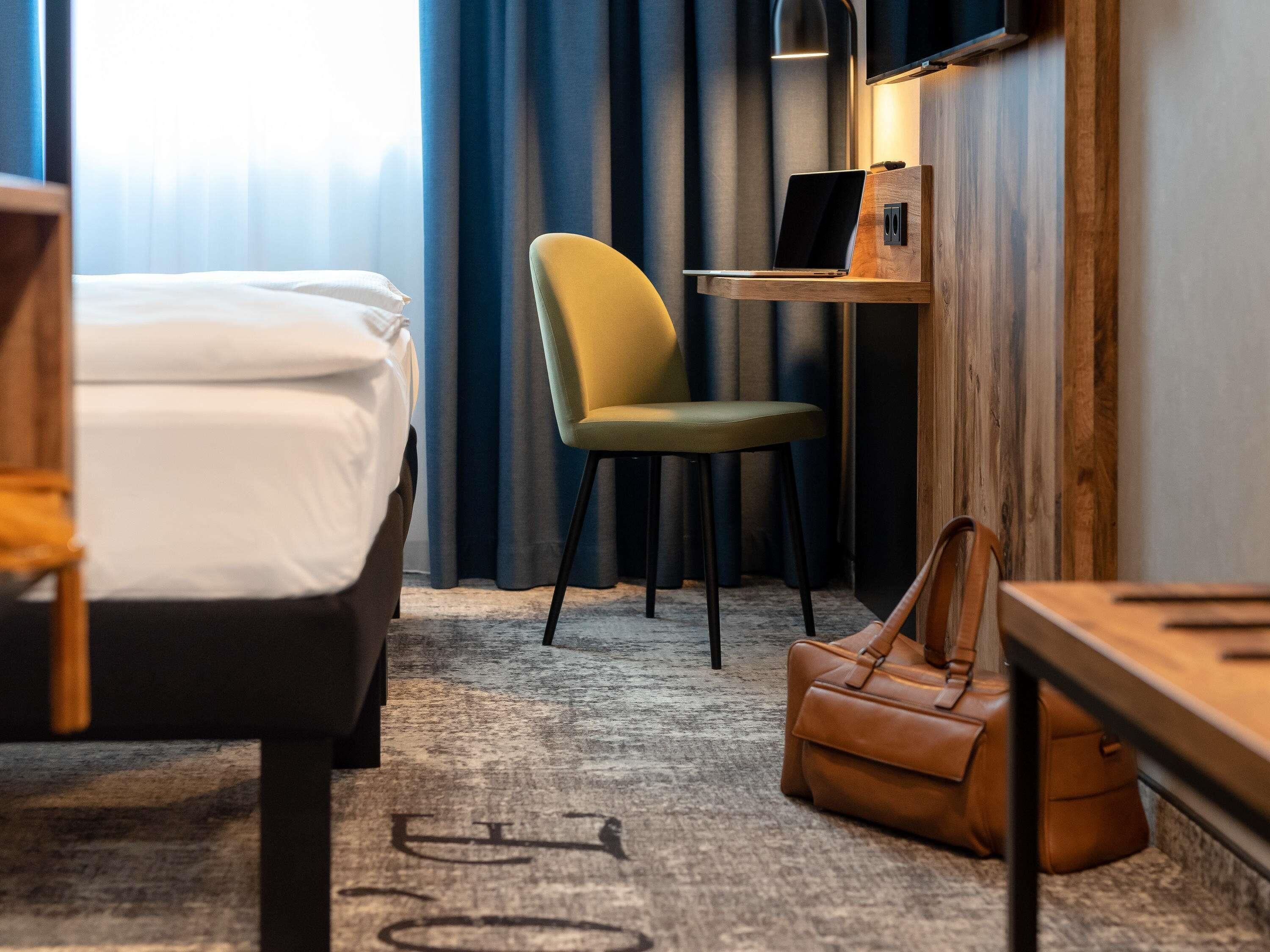Habitación de hotel con cama, una silla, un escritorio con una computadora portátil, un bolso de mano y un ventanal con cortinas.