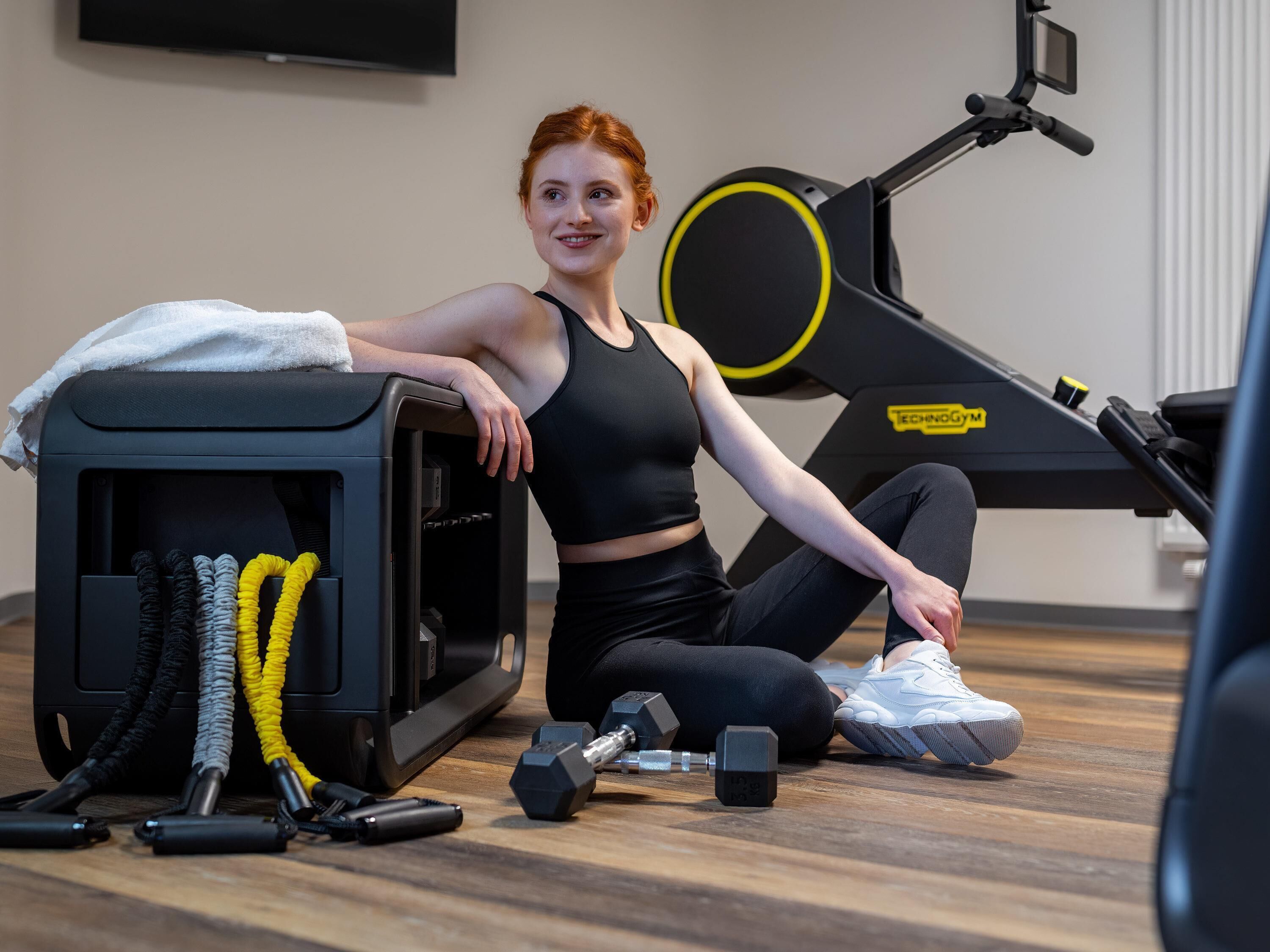A person sitting on the floor next to a stationary bike and a treadmill in a gym.