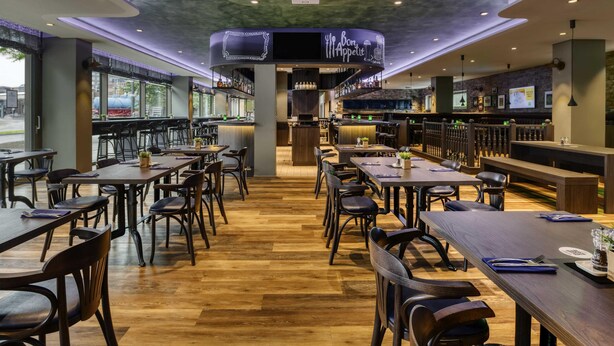 Een restaurant interieur met houten vloeren, donkere tafels en stoelen, een bar en een plafond met inbouwverlichting.