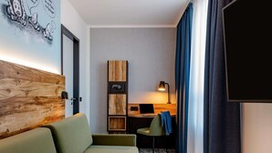 Ein modernes Hotelzimmer mit einem hölzernen Kopfteil, einem Sofa, einem Schreibtisch mit einem Laptop, einem kleinen Stuhl und einem Fernseher.