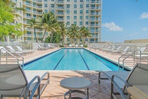 A swimming pool with lounge chairs and a table in the foreground, surrounded by palm trees and a hotel building in the background.
