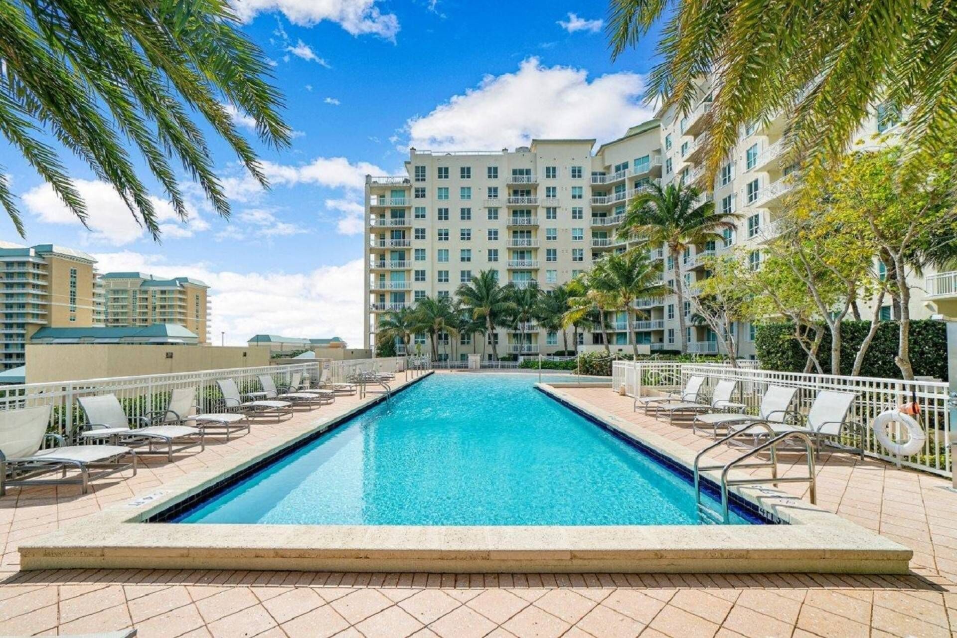 A swimming pool with lounge chairs and palm trees, surrounded by a multi-story building with balconies.